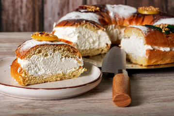A slice of Roscon de Reyes filled with cream on a plate, with the rest of the Roscon in the background, on a wooden table.