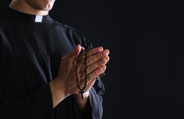 Priest with rosary praying on black background, closeup. Space for text