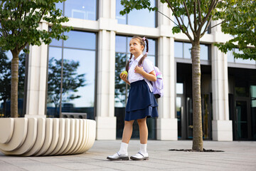 A young girl in school uniform stands outside a modern building, holding an apple and wearing a...