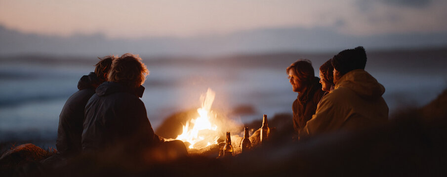 Friends gathered around a bonfire on a serene beach at dusk, sharing stories and enjoying drinks. Warm light, connection, and nature create a peaceful atmosphere.