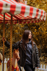 Beautiful happy young brunette girl in a black leather jacket stands near the carousel in the autumn park. autumn concept. autumn season. fall.
