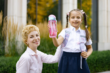 A smiling mother and daughter pose in front of a school building. The girl holds up her water bottle.