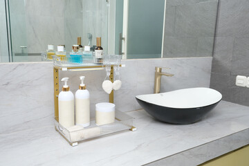 A two-tiered, gold-framed bathroom organizer with bottles of cosmetics is on a marble-patterned countertop next to a black and white basin