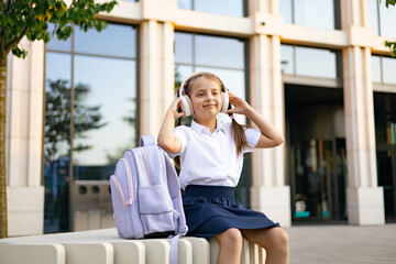 Caucasian schoolgirl wearing headphones, sitting outside the school building.