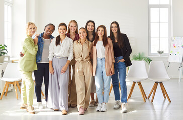 Full length portrait of happy smiling young diverse female company employees wearing casual clothes standing in modern office and looking cheerful at camera. Teamwork and business concept.