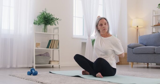 Fit senior woman doing yoga at home. Old gray haired woman sitting on exercise mat with arms behind her back stretching her shoulders and relaxing stiff muscles. Fitness and wellness concept 