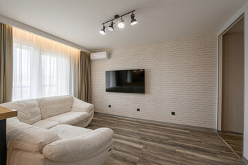 modern living room featuring a beige couch, a flat-screen TV on a brick wall, and wooden flooring....