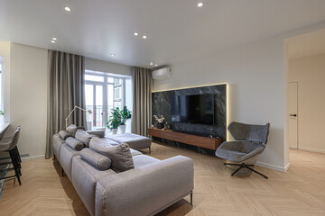 Modern living room with a large sofa, TV wall, and herringbone wood floors. Neutral tones and clean...