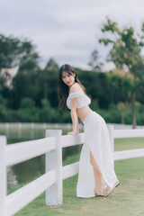 Graceful Beauty in Serene Setting: A young woman, clad in elegant attire, leans casually against a...