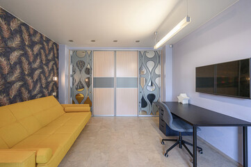 Interior shot of a modern room with a yellow sofa, dark desk, TV, and patterned wardrobe. The walls are a combination of patterned wallpaper and light purple paint