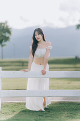Naklejka premium Elegant Pose by Fence: A graceful woman in a flowing white dress leans against a rustic fence, her presence enhanced by a serene outdoor setting.