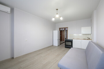 well-lit, compact studio apartment features a minimalist kitchen, pale gray walls, a light wood-look floor, and a simple sofa