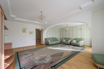 Spacious living room with light walls, wooden floors, and a large patterned rug. Green sofas are...