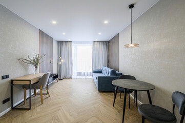 Cozy apartment living room with a blue sofa, dining table, desk, and herringbone floor. Modern...