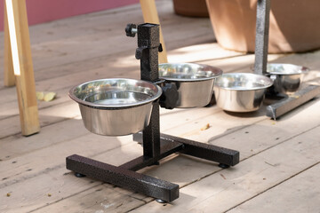 A set of stainless steel pet food bowls on a black adjustable stand, placed on a wooden floor. The background features potted plants and a colorful wall.