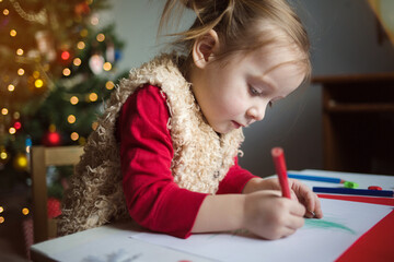 child writes letter to Santa Claus. little girl draws with crayons on background of Christmas tree.