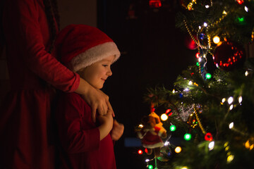 thoughtful child in Santa hat sits in dark near Christmas tree and dreams.