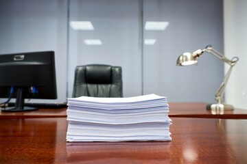 A stack of papers on a wooden desk in an office with a computer and a desk lamp near