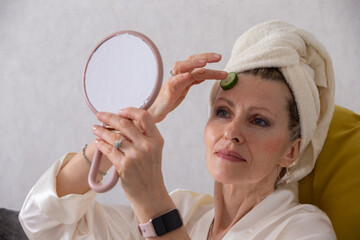 Woman applying cucumber slice on forehead while looking in mirror at home during self-care routine
