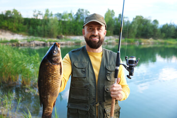 Smiling fisherman holding rod and caught fish near lake at summer