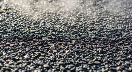 Close-up of rough dark asphalt or gravel texture with shimmering light and shallow depth of field, showing granular surface detail.