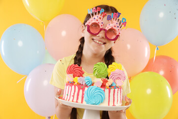 Birthday celebration. Cute little girl with tasty and balloons on orange background