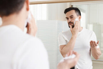 Handsome man applying shaving foam near mirror in bathroom