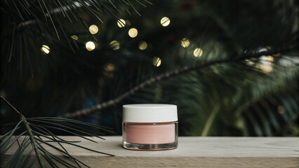 pink cosmetic cream jar with a white lid, on a wooden surface with a christmas tree and festive lights in the background