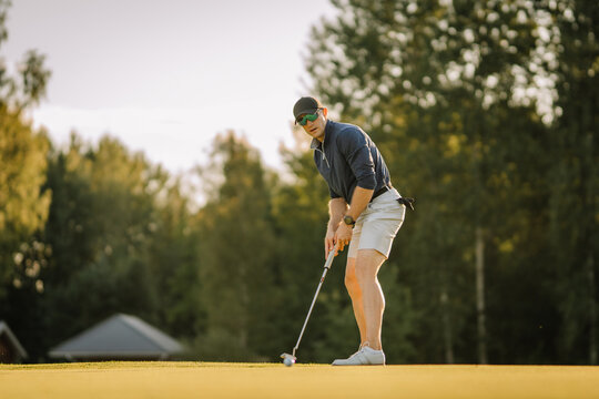 Focused male golfer prepares to putt on a sunny green, eyes locked on the ball. Surrounded by forest trees and evening light during a peaceful game.