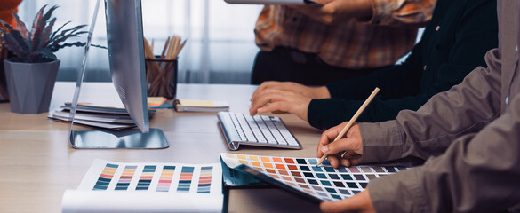 A diverse group of professionals engaging in a creative brainstorming session in a modern office. Color palettes, a computer, and various tools lay on the wooden desk, fostering inspiration. SACTR