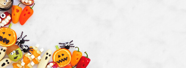 Healthy Halloween fruit and vegetable snacks. Assortment of fun, spooky treats. Overhead view corner border over a white marble banner background with copy space.