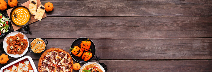 Spooky Halloween dinner party corner border over a dark wood banner background. Overhead view. Skull pizza, salad, pasta, ghost lasagna, mummy pot pie, pumpkin soup, jack o lantern stuffed peppers.