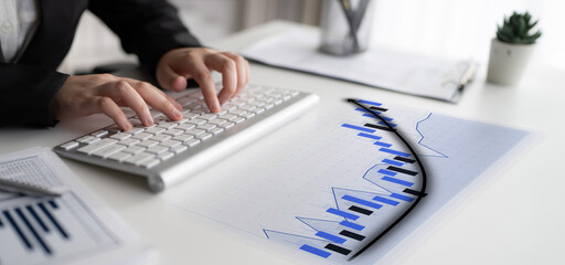 A business professional focuses on financial data using a keyboard, surrounded by charts and documents on a modern office desk, fostering productivity and analysis. Impute
