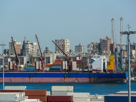 Alexandria, Egypt - 05.04.2025  Container cargo ship with loading cranes and stacked shipping containers at the Port of Alexandria, Egypt, with city skyline in the background.