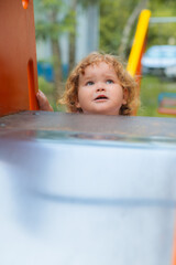 A young child with curly hair gazes curiously at a bright play structure in a bustling park.