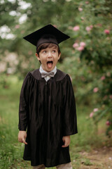 Portrait of emotional boy-graduate in graduation dress and hat on green background, spring. Children's education concept