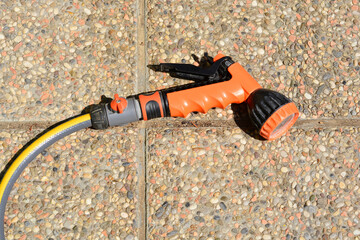 An orange and black garden hose nozzle lies on a textured stone patio, with a distinct shadow cast...
