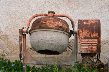 An old, rusty concrete mixer stands in front of a wall, surrounded by overgrown weeds and wild...