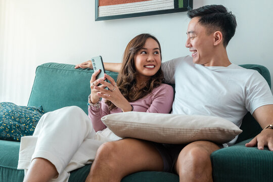 Smiling young Asian couple sitting on green sofa at home, looking at each other while holding smartphone. Woman in pink top, man in white t-shirt with smartwatch, cozy indoor setting