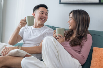 Fototapeta premium Smiling young Asian couple sitting on green sofa at home, enjoying hot drinks together. Woman in pink top, man in white t-shirt with smartwatch, cozy indoor living room background