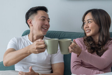 Smiling young Asian couple sitting on green sofa at home, toasting coffee mugs together. Woman in pink top, man in white t-shirt with smartwatch, cozy indoor living room background