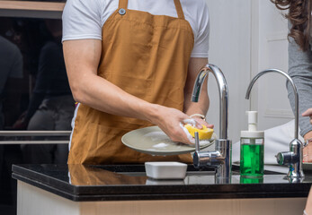 Asian man wearing a brown apron washing dishes with a sponge and soap at the kitchen sink,...