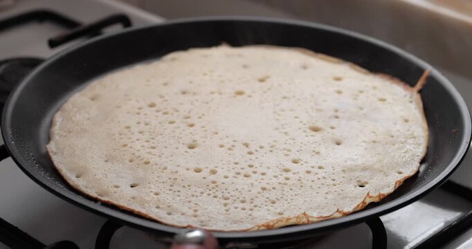 A thin pancake cooking in a black frying pan on a gas stove. Bubbles form on the surface as it cooks evenly.