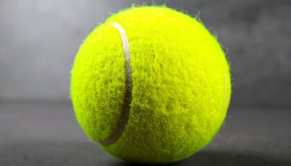 A vibrant yellow tennis ball rests on a muted gray surface, showcasing its textured surface and the prominent white outlining.