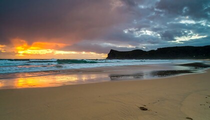 Dramatic sunrise paints a golden hue across the beach, with waves crashing against the shore and a dark cliff rising majestically in the distance.