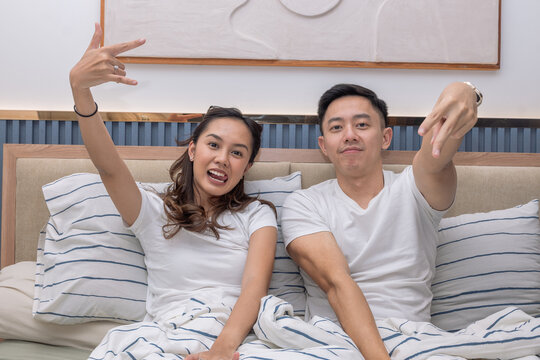 Asian couple sitting on bed playfully making hand signs and smiling, showing fun and relaxed energy in a cozy bedroom with striped bedding - Powered by Adobe