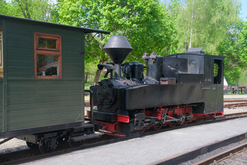 historic narrow gauge steam locomotive waits at forest station in bright spring light – black iron engine meets green wooden carriage beneath fresh trees and blue sky