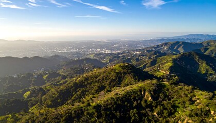 Naklejka premium Expansive Aerial View of a Sunlit Cityscape Nestled Among Lush Green Hills Under a Clear Blue Sky.