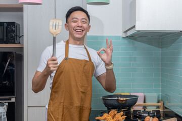 Asian man in apron smiling in the kitchen, holding a spatula and making an OK hand gesture, standing near stove with frying pan, fried chicken, and eggs on the counter