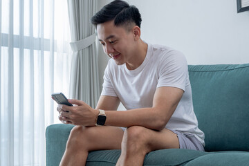 Young Asian man sitting on green sofa near window with curtains, smiling while holding smartphone....